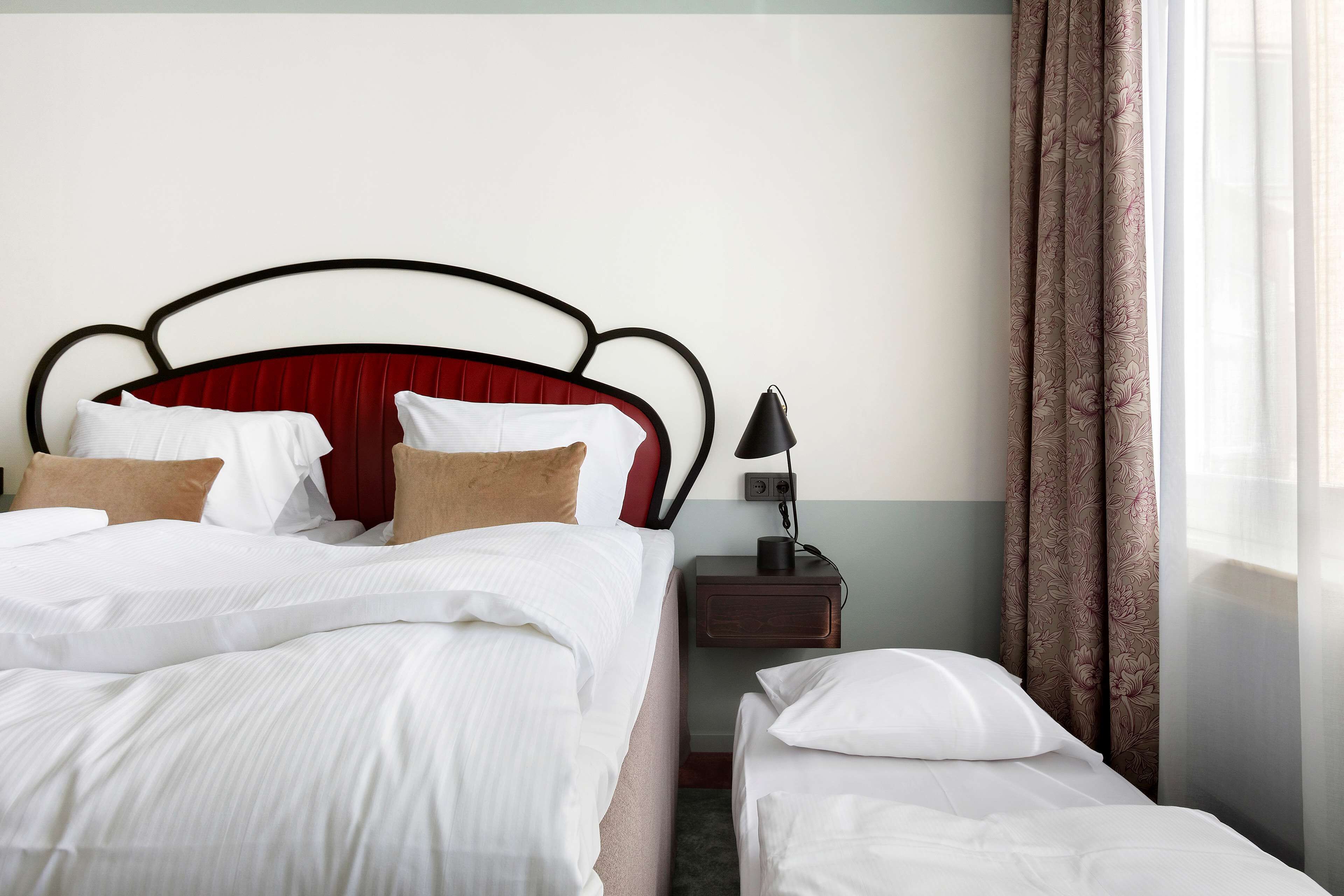 A hotel room with a double bed, a single bed, a nightstand with a lamp, and a window with curtains.