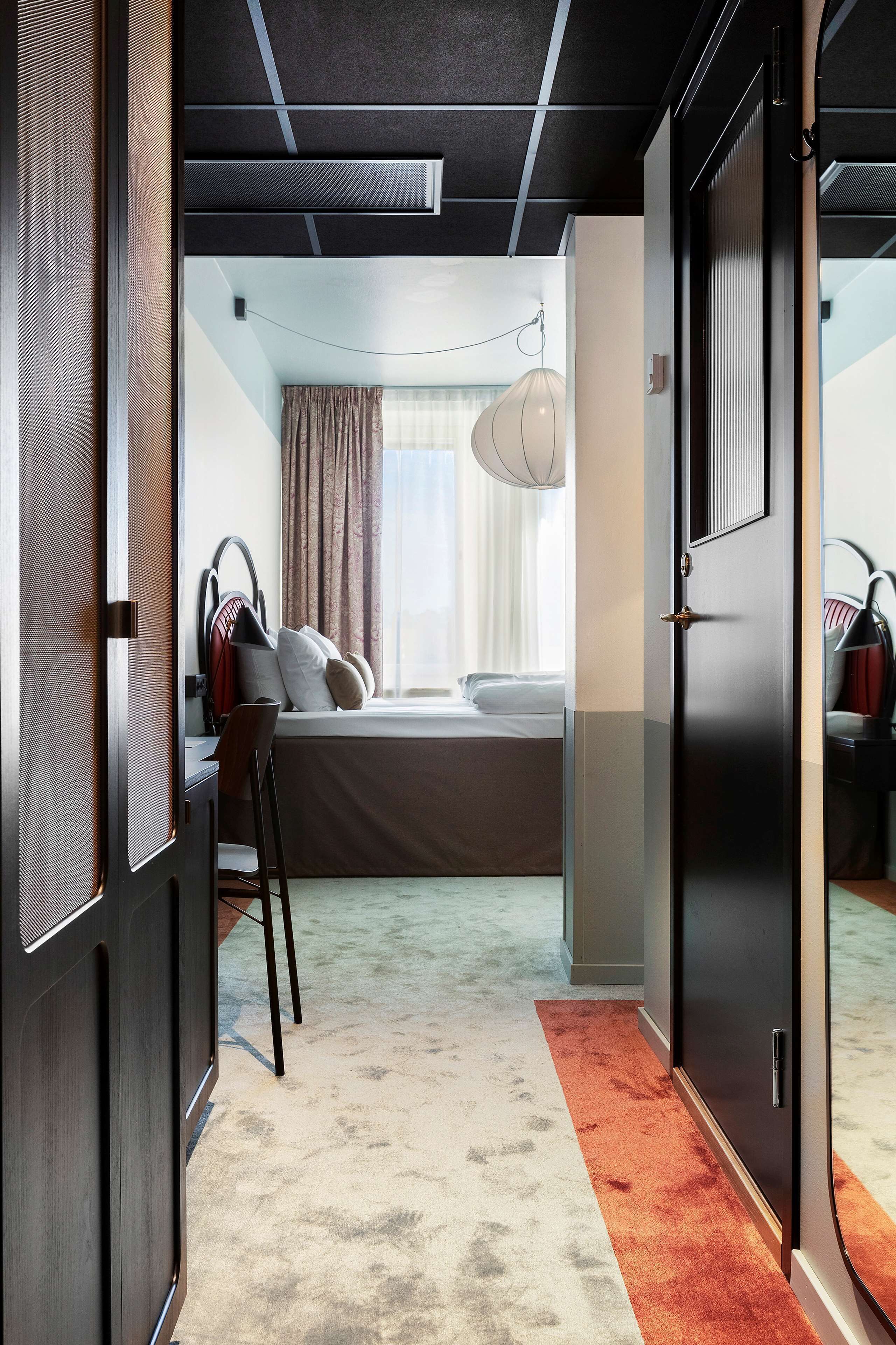 Ett hotellrum med en säng, ett skrivbord, en stol, en garderob och ett fönster med gardiner.