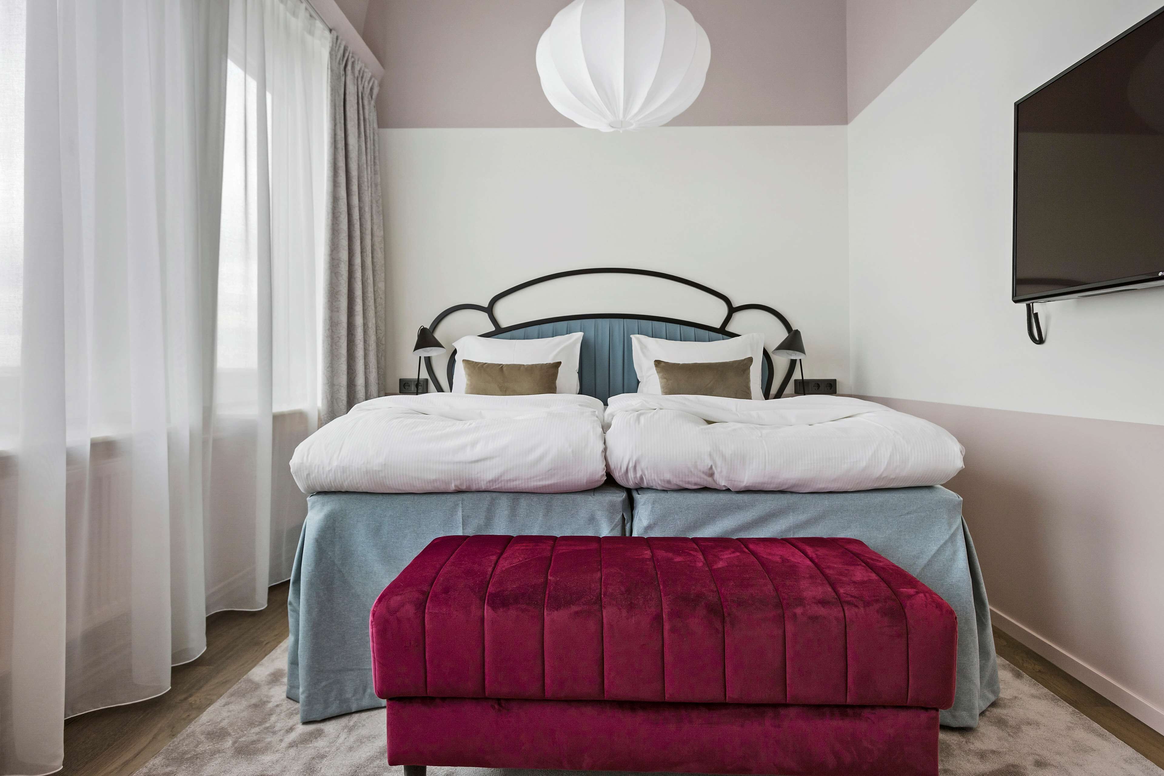 A bedroom with a bed, a red ottoman, a TV, and a window with curtains.