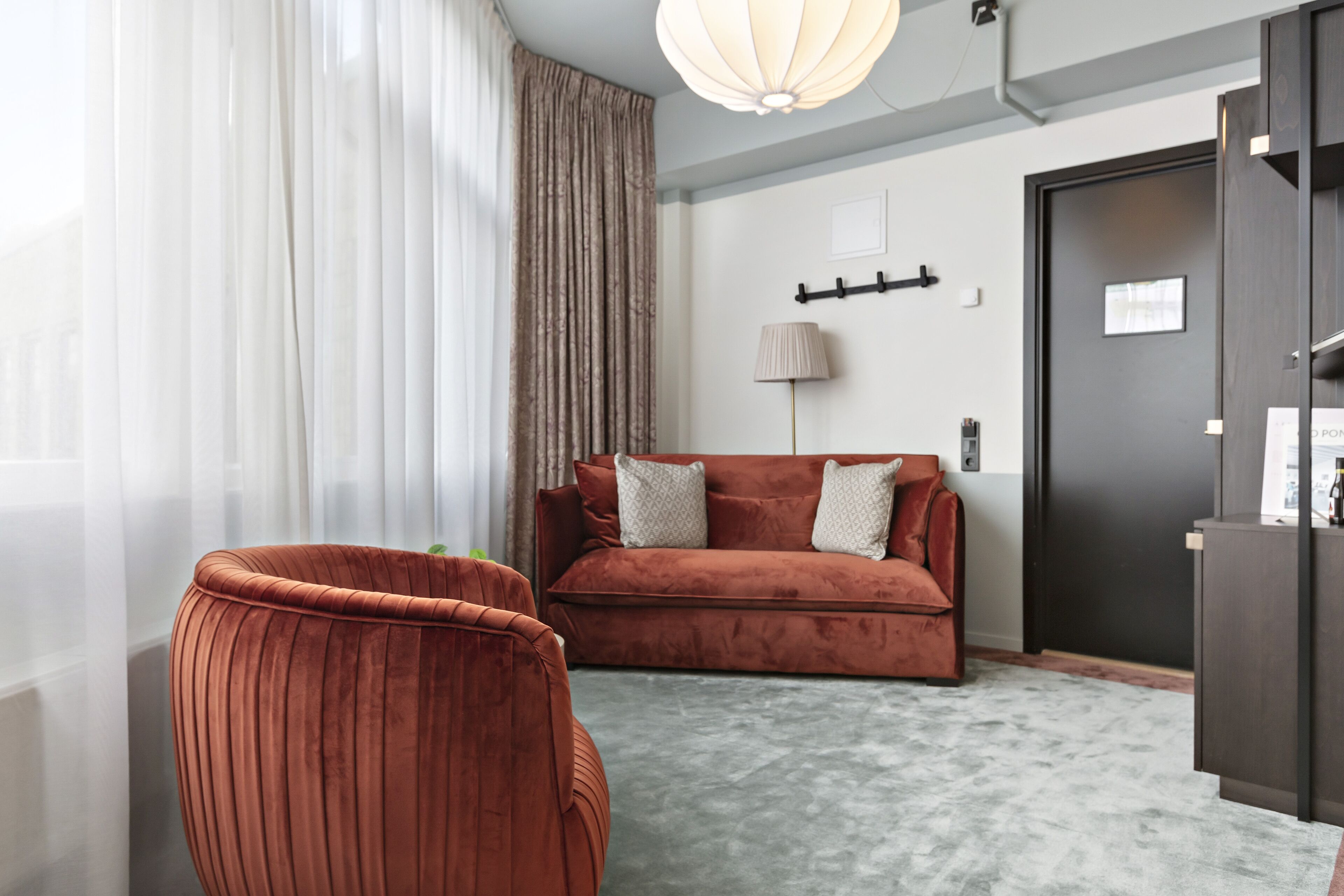 A modern living room with a red sofa, a grey carpet, and a large window with sheer curtains.