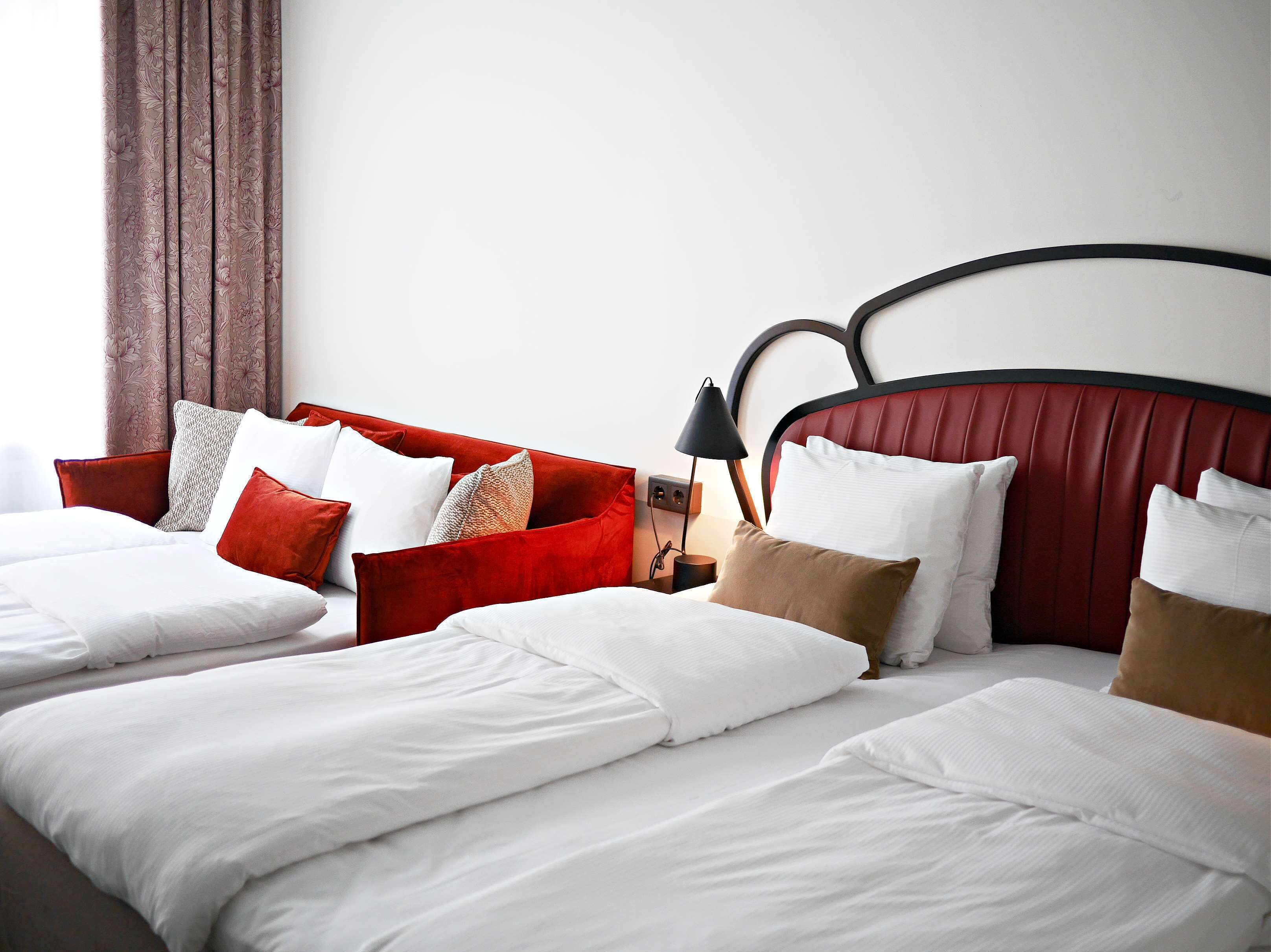 Habitación de hotel con cama doble, cabecera tapizada de rojo y mesita de noche con lámpara.