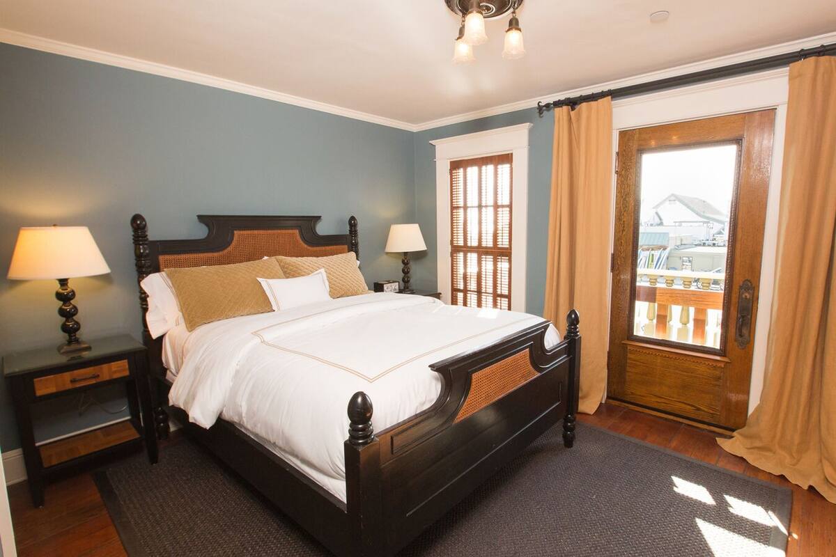 A neatly made bed with a wooden headboard, two bedside tables with lamps, a window with curtains, and a view of a building outside.