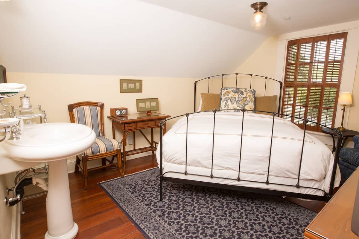 A bedroom with a bed, a bedside table, a chair, a desk, a lamp, and a window with blinds.