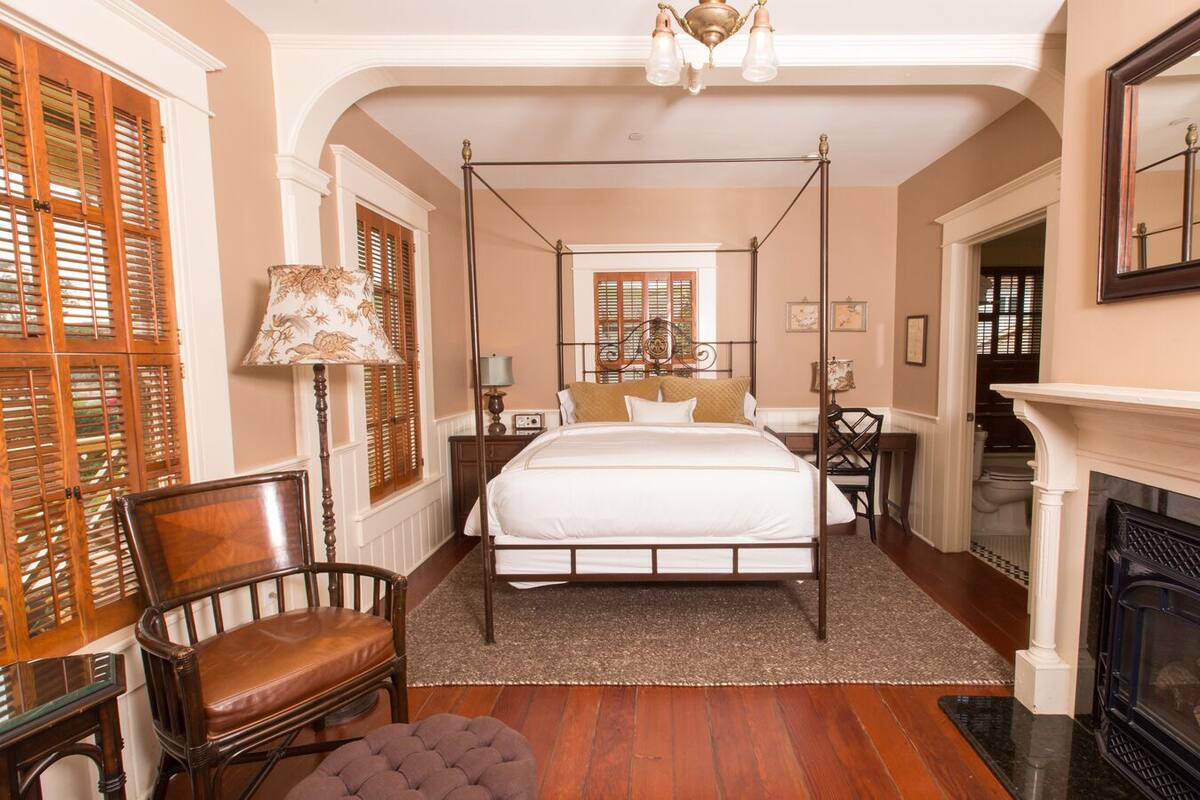 A four-poster bed with a white bedspread, a wooden chair, a small ottoman, a fireplace, and a mirror on the wall.