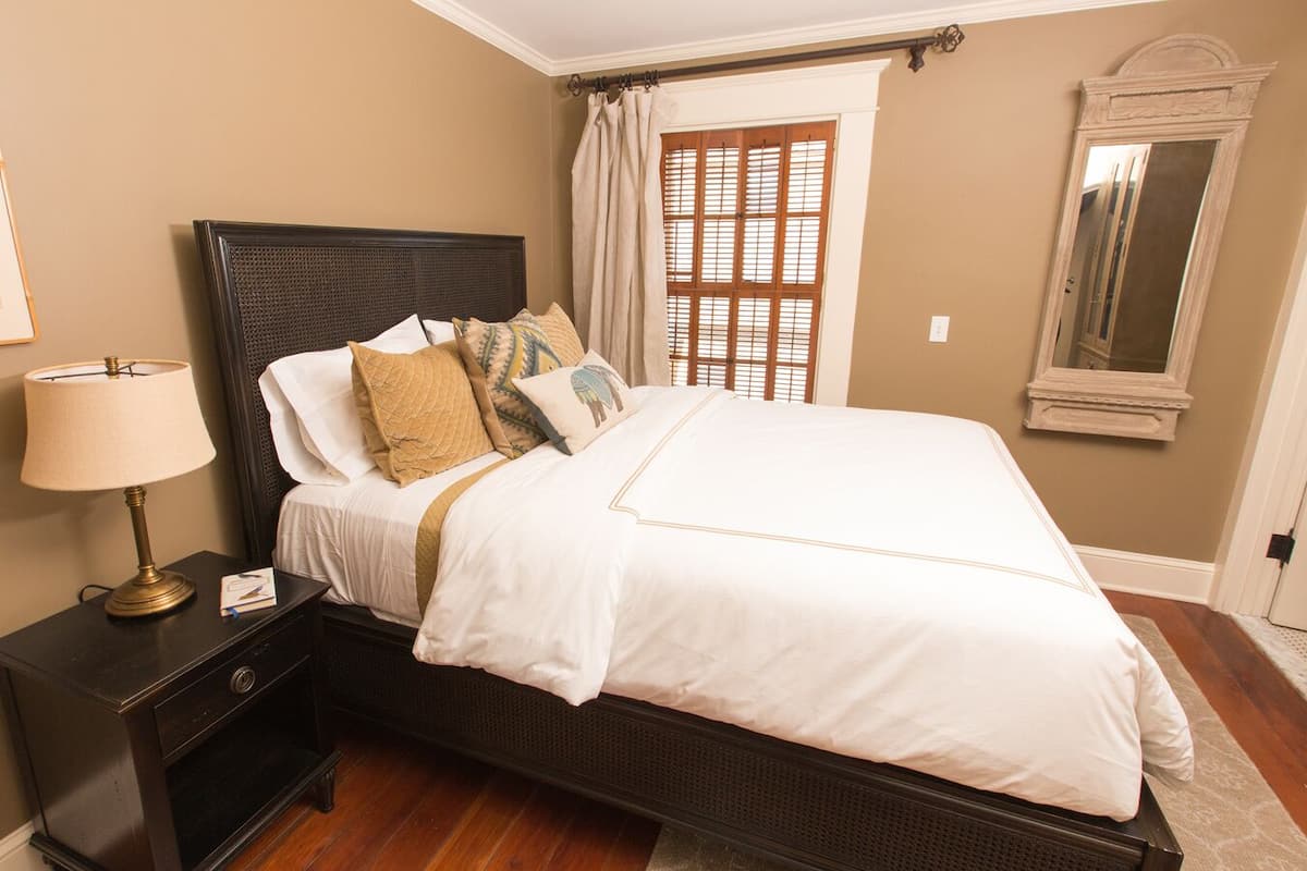 A neatly made bed with a white comforter and multiple pillows, a bedside table with a lamp, a framed picture on the wall, and a window with wooden shutters.