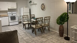 A compact kitchen with white cabinets, a white refrigerator, a stove, and a dining area with a glass table and wooden chairs.