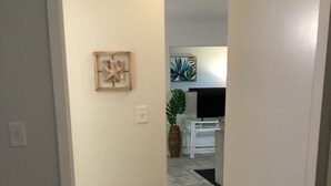 A hallway leading to a living area with a flat-screen TV, a plant, and a framed picture on the wall.