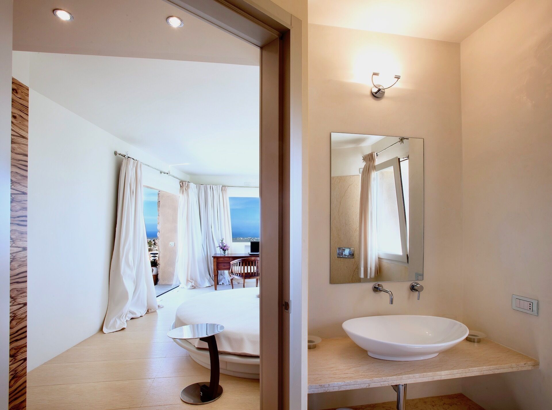 Ein modernes Hotelzimmer mit einem großen Bett, einem Nachttisch, einem Badezimmer mit Waschbecken und Spiegel sowie Blick aufs Meer.