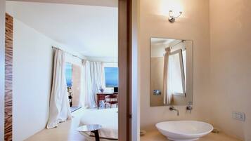 Ein modernes Hotelzimmer mit einem großen Bett, einem Nachttisch, einem Badezimmer mit Waschbecken und Spiegel sowie Blick aufs Meer.