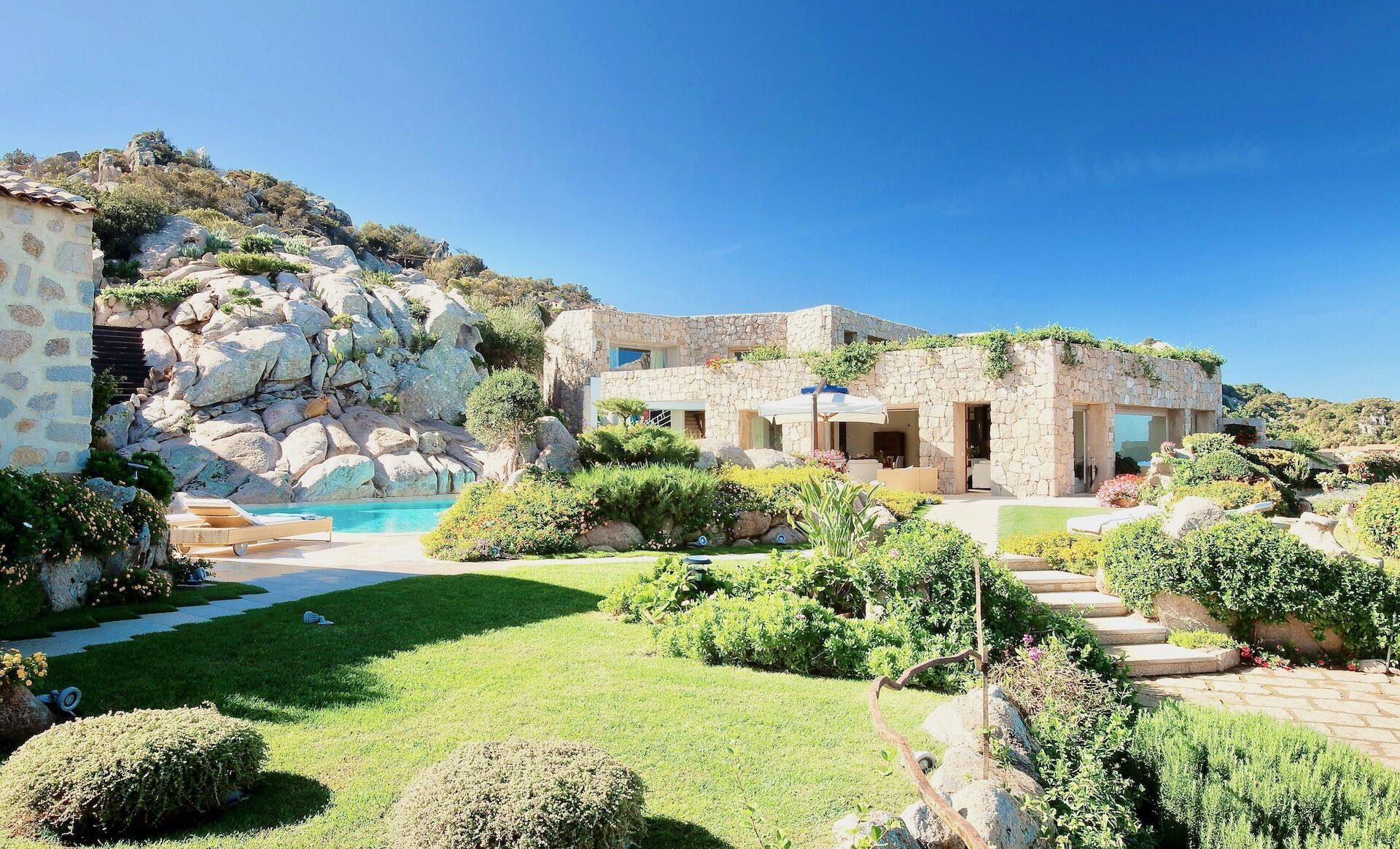 Una villa de piedra con piscina, rodeada de exuberante vegetación y un terreno montañoso rocoso.