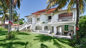 A two-story building with a red-tiled roof, white walls, and a green flag. There is a staircase leading to the upper floor, and the building is surrounded by a well-maintained lawn and palm trees. - Casablanca Curral Beach (ilhabela)