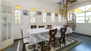 A dining area with a table set for six, surrounded by wooden chairs with heart-shaped cutouts. There are six arched windows with yellow-tinted glass, and a spiral staircase with golden railings is visible in the background. - Casablanca Curral Beach (ilhabela)