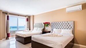 Hotellihuone, jossa on kaksi sänkyä, suuri ikkuna, jossa on verhot, ilmastointilaite ja näkymä merelle.