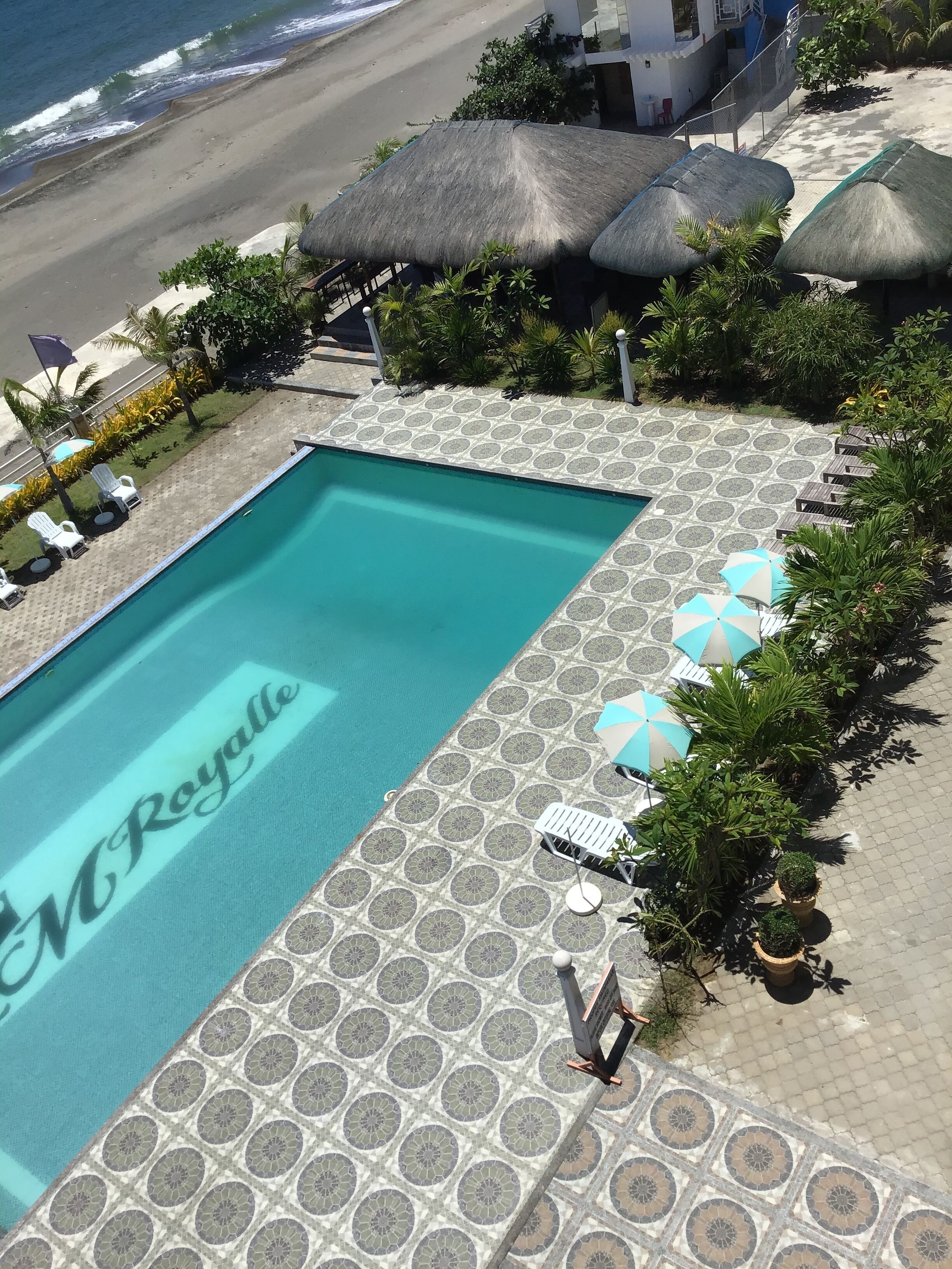 Ein Pool mit einem gemusterten Fliesenbelag, Liegestühlen, Sonnenschirmen und Blick auf den Strand.
