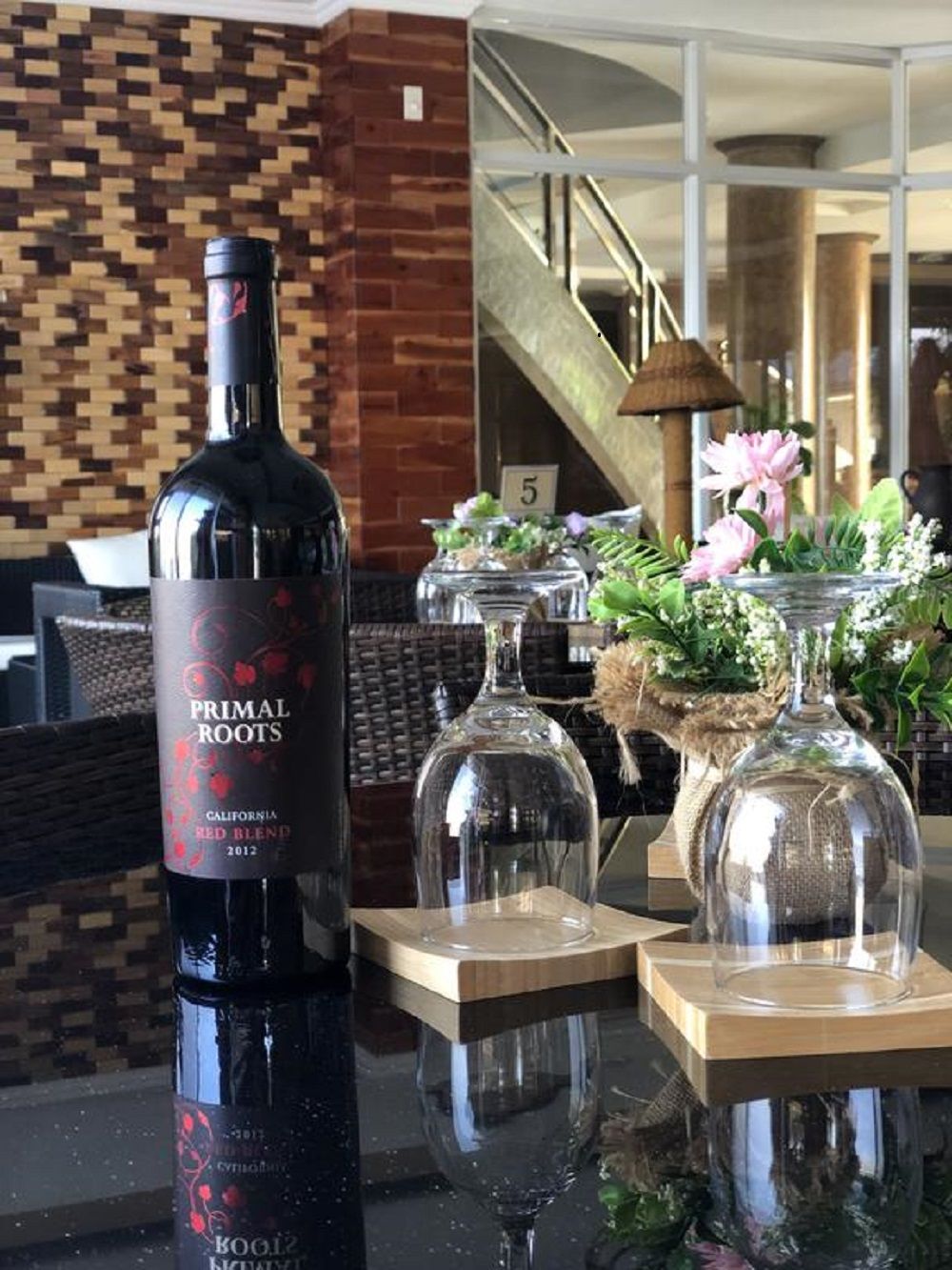 A bottle of Primal Roots California Red Blend wine, a glass decanter, and a vase with flowers on a table.