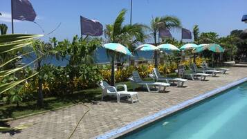 A poolside area with lounge chairs and umbrellas, surrounded by palm trees and a clear sky.
