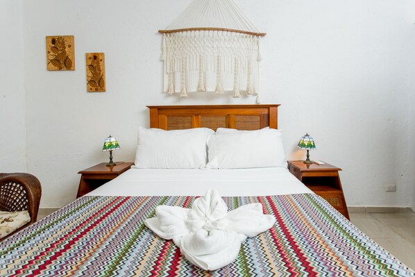 A neatly made bed with a colorful blanket, a white towel, and a macramé hanging above. There are two bedside lamps and a small wooden nightstand with a lamp. - Aldea JOYAH (Tulum)