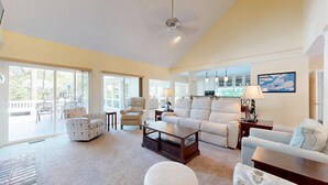 A spacious living room with a large sofa, a coffee table, and a dining table. There are large windows providing natural light and a view of the outdoors. - Very Nice Home Located on the 11-Mile Lagoon with Its Own Large Private Pool! (Hilton Head)