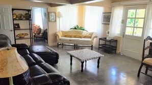 Una sala de estar con un sofá, una mesa de centro y un sillón. Hay una puerta con ventanal, una lámpara y una estantería con adornos.
