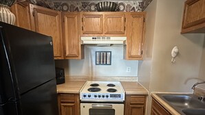 Cocina compacta con armarios de madera, un refrigerador negro, una estufa de cuatro quemadores y un lavamanos.