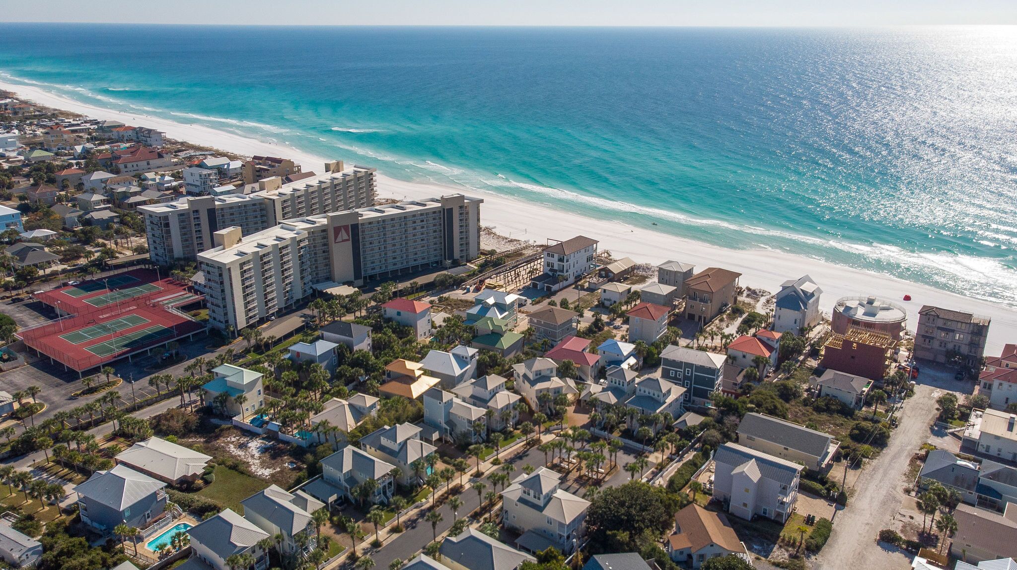 An aerial view of a coastal residential area with beachfront properties, a large hotel complex, and a tennis court facility.