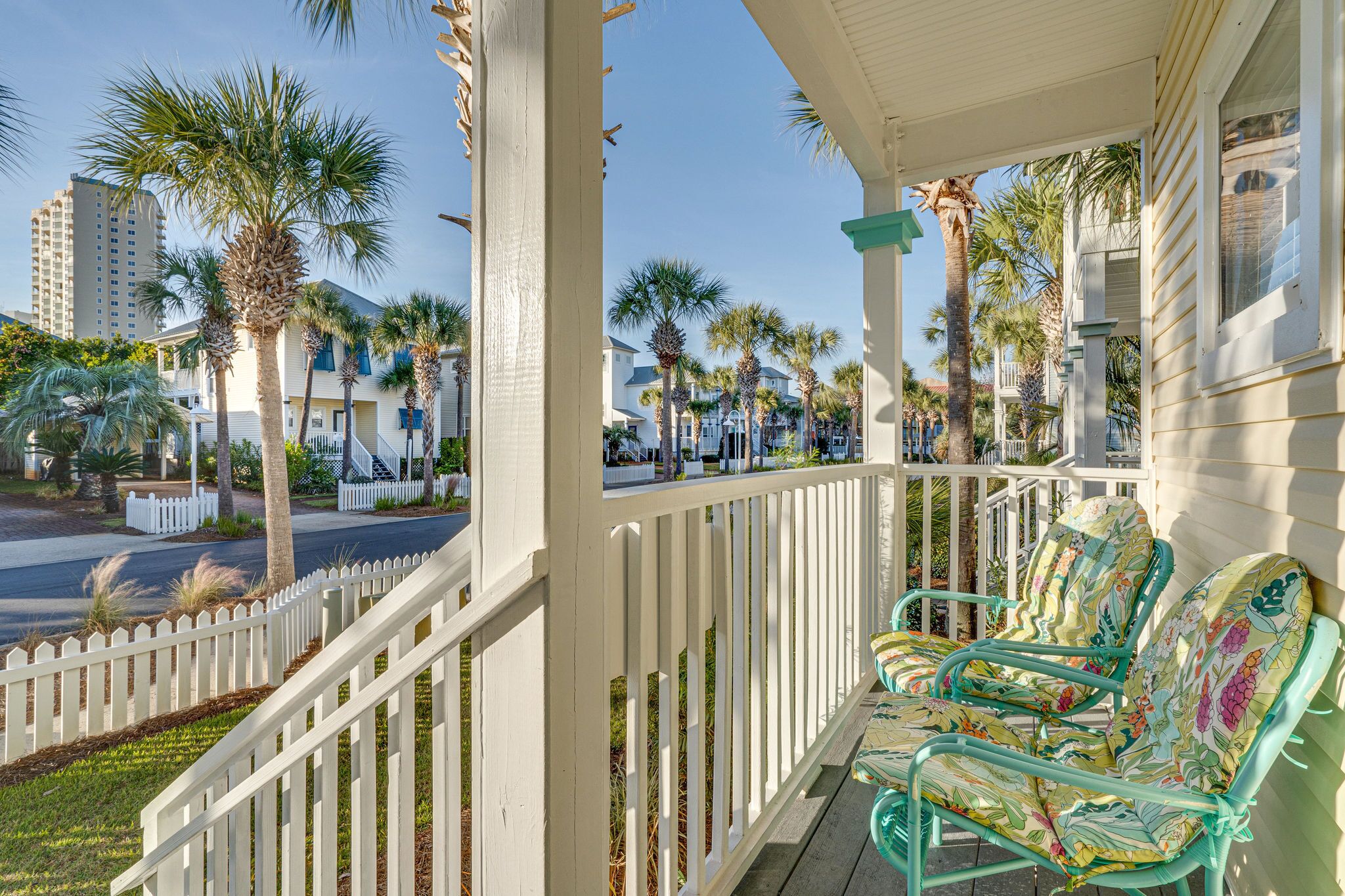 A porch with white picket fence, green metal chairs with floral cushions, and palm trees in the background.