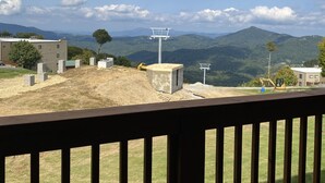 Vista desde un balcón con baranda de madera, sobre un sitio de construcción con un edificio en construcción, una excavadora amarilla y un cielo despejado con nubes.