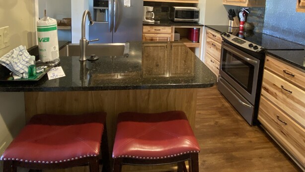 Una cocina con gabinetes de madera, un refrigerador de acero inoxidable, un microondas y una estufa. Hay dos taburetes rojos en la isla.