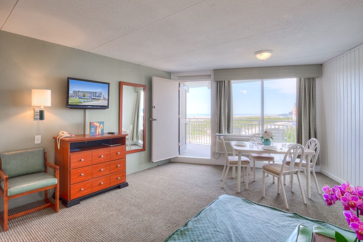 Een hotelkamer met een bed, een bureau, een dressoir, een stoel, een eettafel met stoelen, een televisie, een raam met uitzicht op het strand en een balkon.