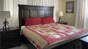 Un lit bien fait, recouvert d’une couette rouge et beige, avec une tête de lit en bois foncé et deux tables de chevet dotées de lampes.