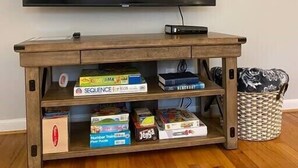 A wooden TV stand with a flat-screen TV mounted above it. The stand has multiple shelves with various board games and a woven basket. - St. Elmo Abode-3 mi to Ruby Falls (Chattanooga)