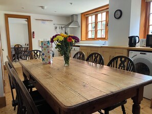Un comedor con mesa de madera, sillas, un florero con flores, microondas, lavadora y ventana con vistas.