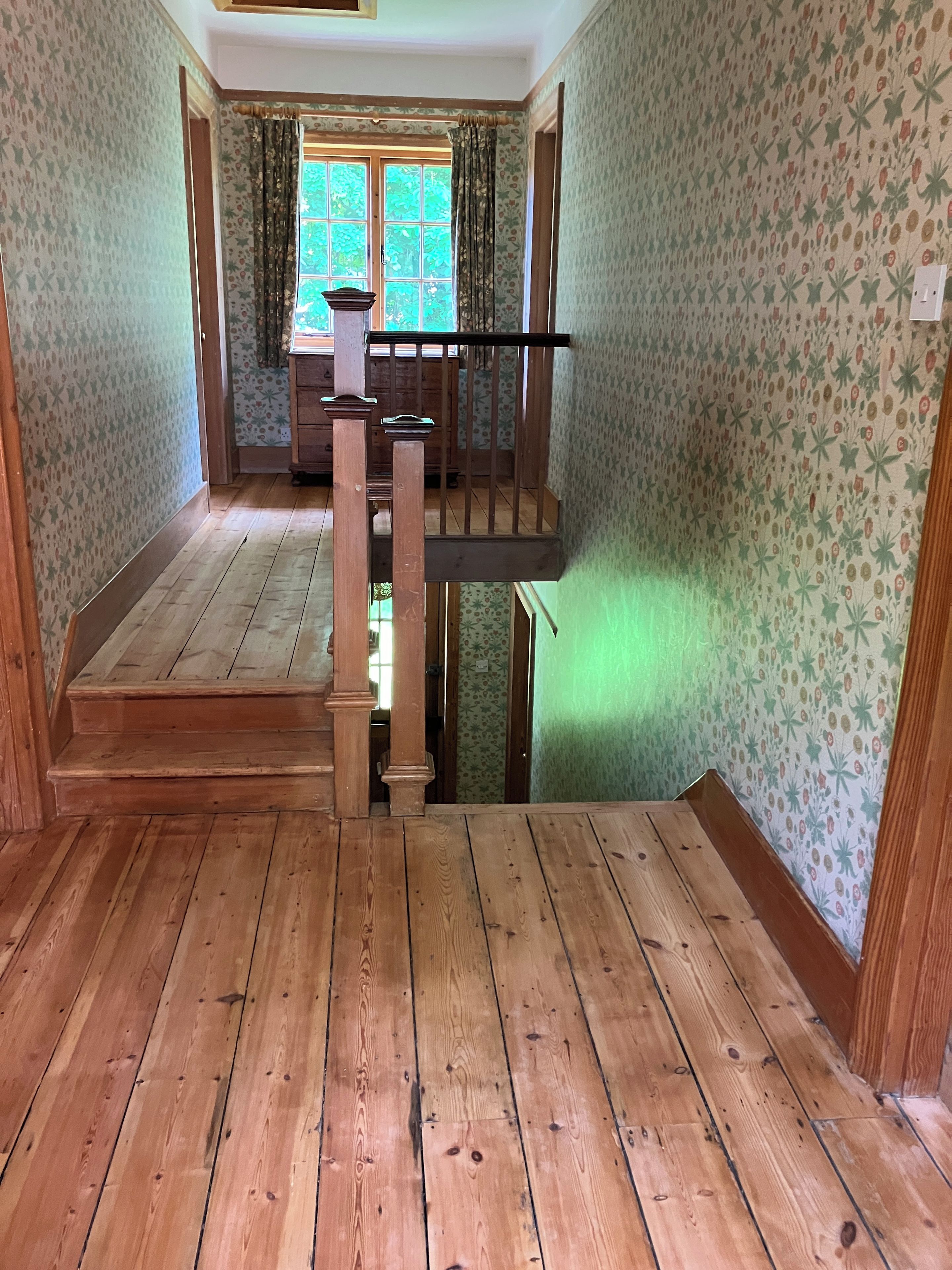A wooden staircase leading to a balcony with floral wallpaper and a window with curtains.