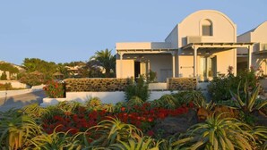 A modern building with a stone facade, a curved roof, and a garden with various plants and flowers. - Alafouzos Studios (Santorini)