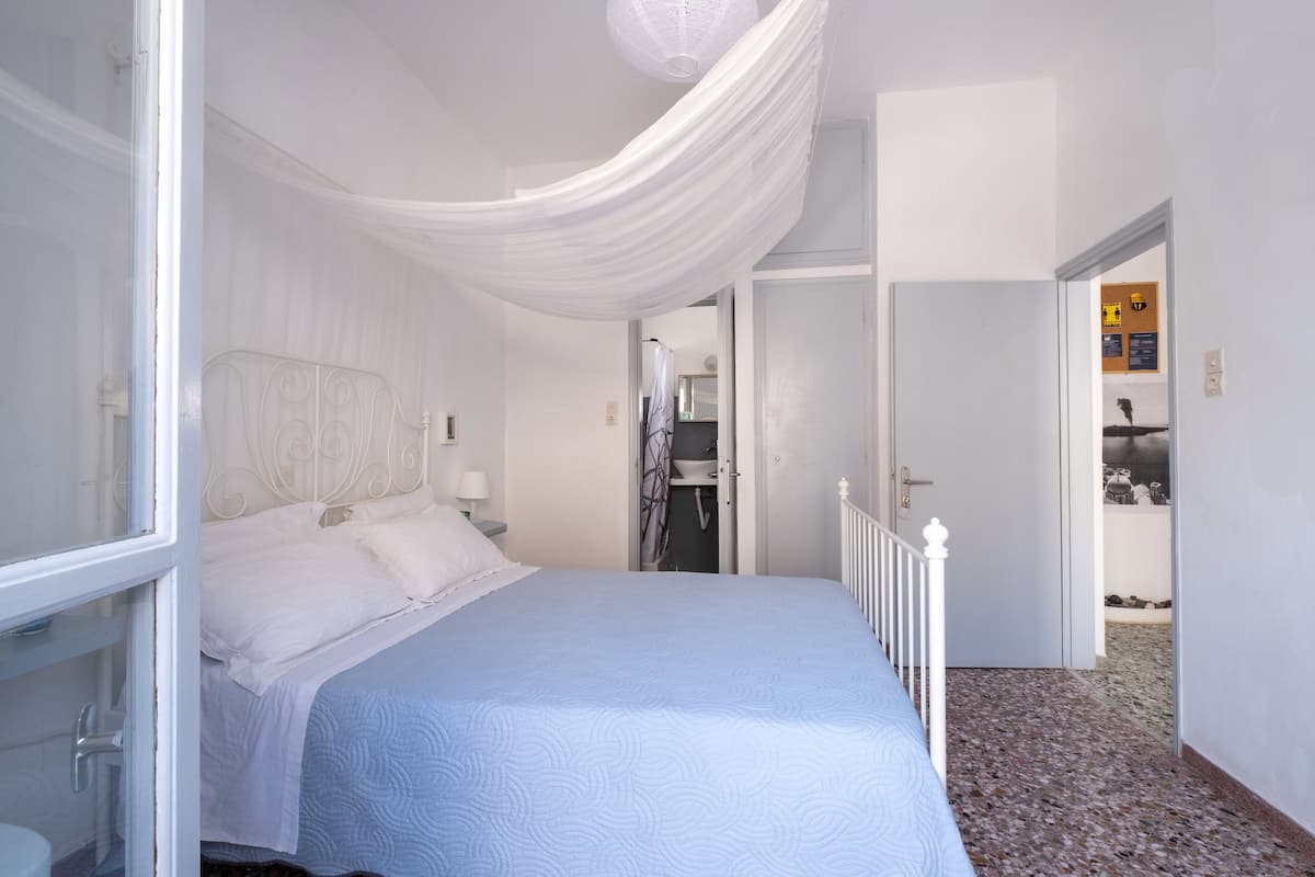 A bedroom with a bed, white curtains, a nightstand, and a door leading to another room.