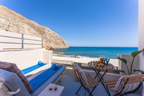 A balcony with a view of the sea, lounge chairs, a table with snacks, and a bottle of water. - Sagma Beach Rooms (Santorini)