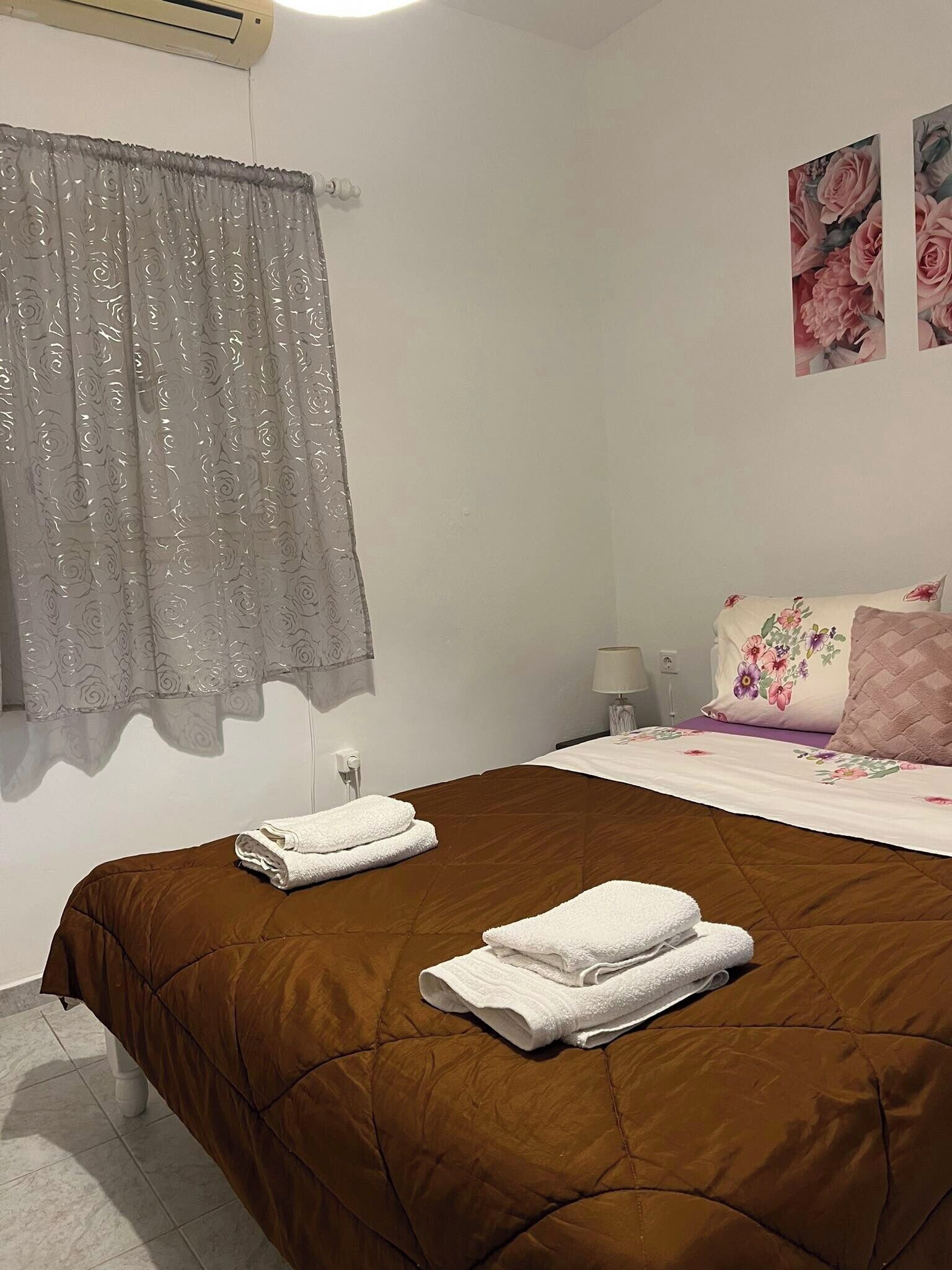 Un dormitorio con colcha marrón, dos toallas blancas dobladas con cuidado, una ventana con cortinas traslúcidas y un cuadro floral en la pared.
