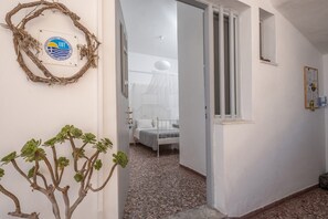A hallway with a bed, a white wall, and a door leading to a bedroom. - Sagma Beach Rooms (Santorini)