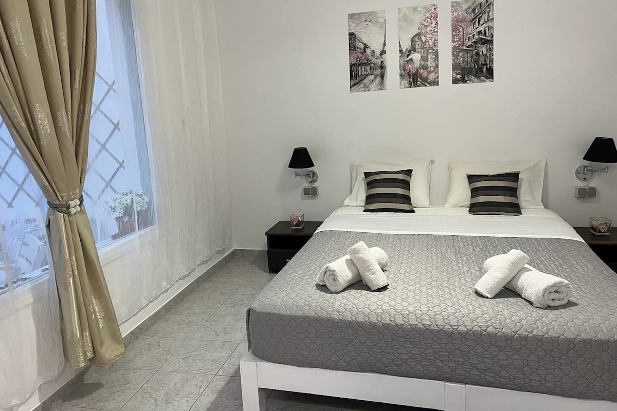 A hotel room with a bed, two bedside tables, a window with curtains, and three wall-mounted pictures.