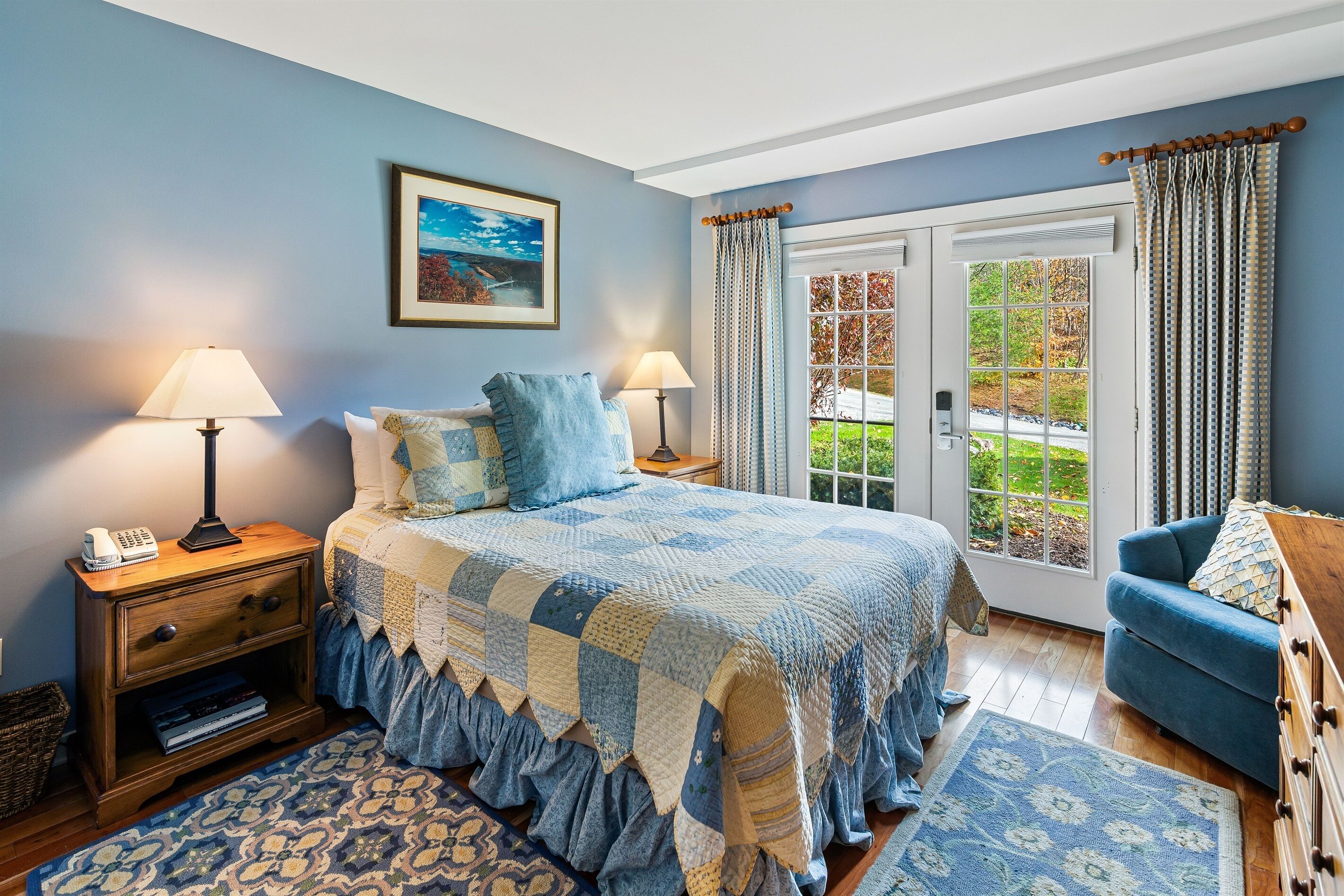 A bedroom with a large bed, two bedside tables with lamps, a blue chair, and a view of the outdoors through a glass door.