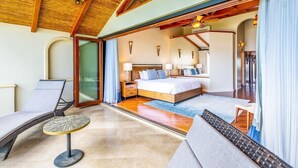 A spacious bedroom with a large bed, a sitting area with a small table, and a view through a glass door to another room.