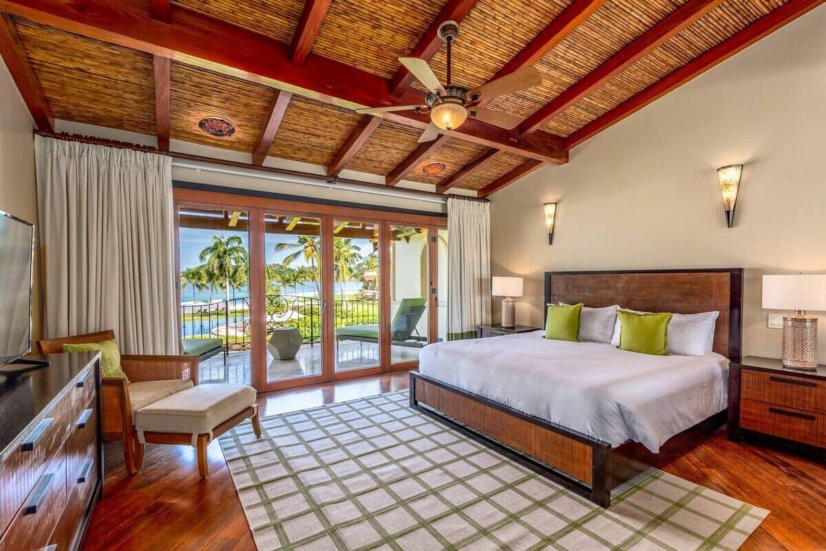 A spacious bedroom with a large bed, a desk, and a chair. There is a balcony with a view of palm trees.