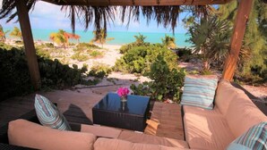 A covered outdoor seating area with a wicker sofa and a table, overlooking a beach with palm trees and clear blue water.