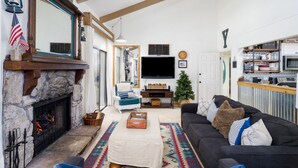 Ein Wohnzimmer mit Kamin aus Stein, einem großen Flachbildfernseher, einem dunkelgrauen Sofa und einer Küchenzeile mit Pizza-Station.