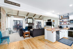 Een ruime woonkamer met een open haard, een bank, een eettafel en een keuken met witte kastjes en een houten aanrechtblad.