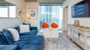 Un salon moderne avec un canapé bleu, un pouf blanc et un tapis à longues mèches gris. Un téléviseur est fixé au mur et une fenêtre offre une vue sur des immeubles de grande hauteur.