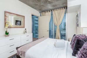 Een moderne hotelkamer met een groot bed, een witte dressoir, een spiegel en uitzicht op een hoog gebouw door een glazen deur.