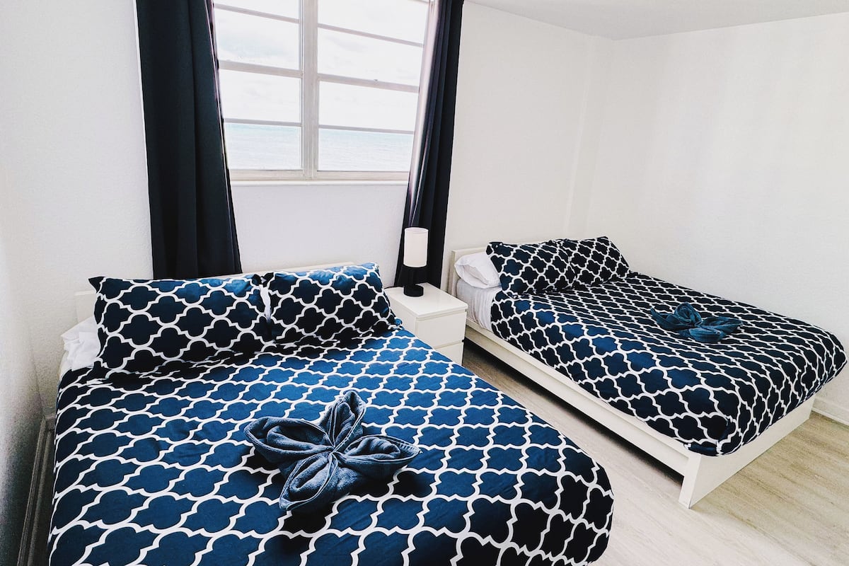 A room with two beds, each with a patterned bedspread and matching pillows. There is a window with curtains, a bedside table with a lamp, and a wall-mounted shelf.