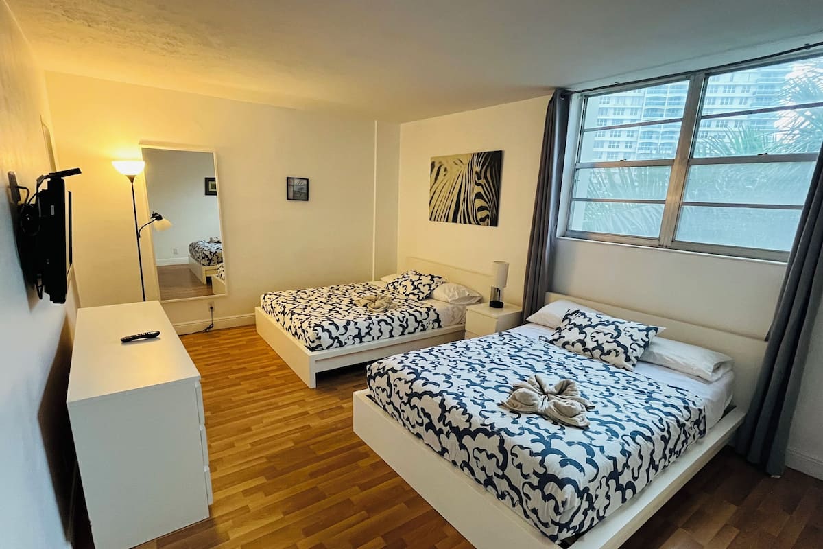 A hotel room with two beds, a desk, a mirror, and a window with a view of buildings.