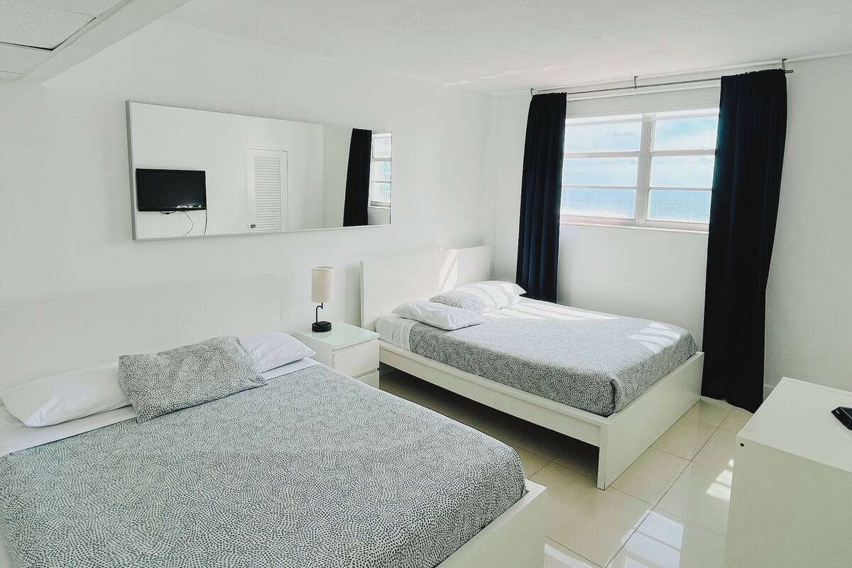 A modern hotel room with two beds, a large window with curtains, a flat-screen TV, and a bedside table with a lamp.